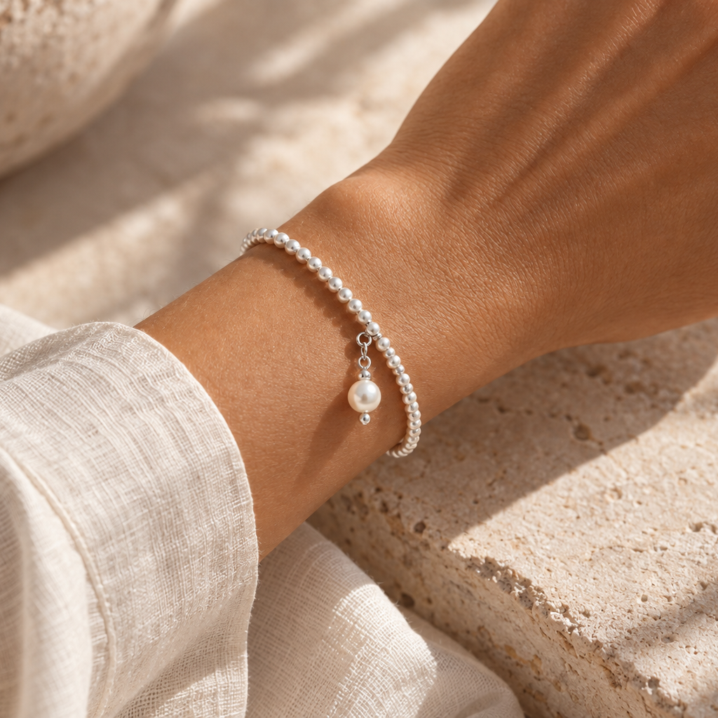 A delicate silver chain bracelet with a single white pearl charm, worn on a person's wrist. The person is wearing a light beige linen long-sleeve top and resting their hand on a textured stone surface under warm, natural sunlight.